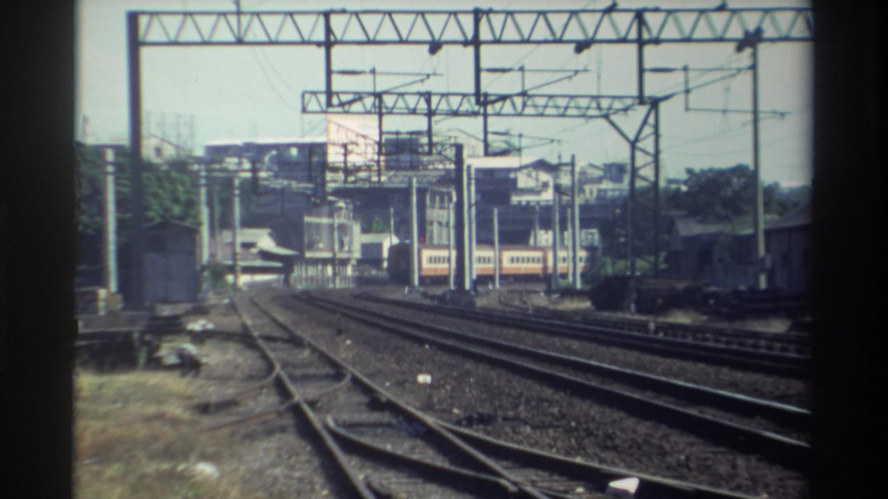 Parallel railroad tracks under overhead electrical gantries with a distant train and rail-side buildings.