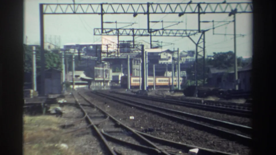 1982 Taiwan home-movie frame showing multiple railroad tracks, overhead electric lines, a distant train, and low buildings beyond the rails.