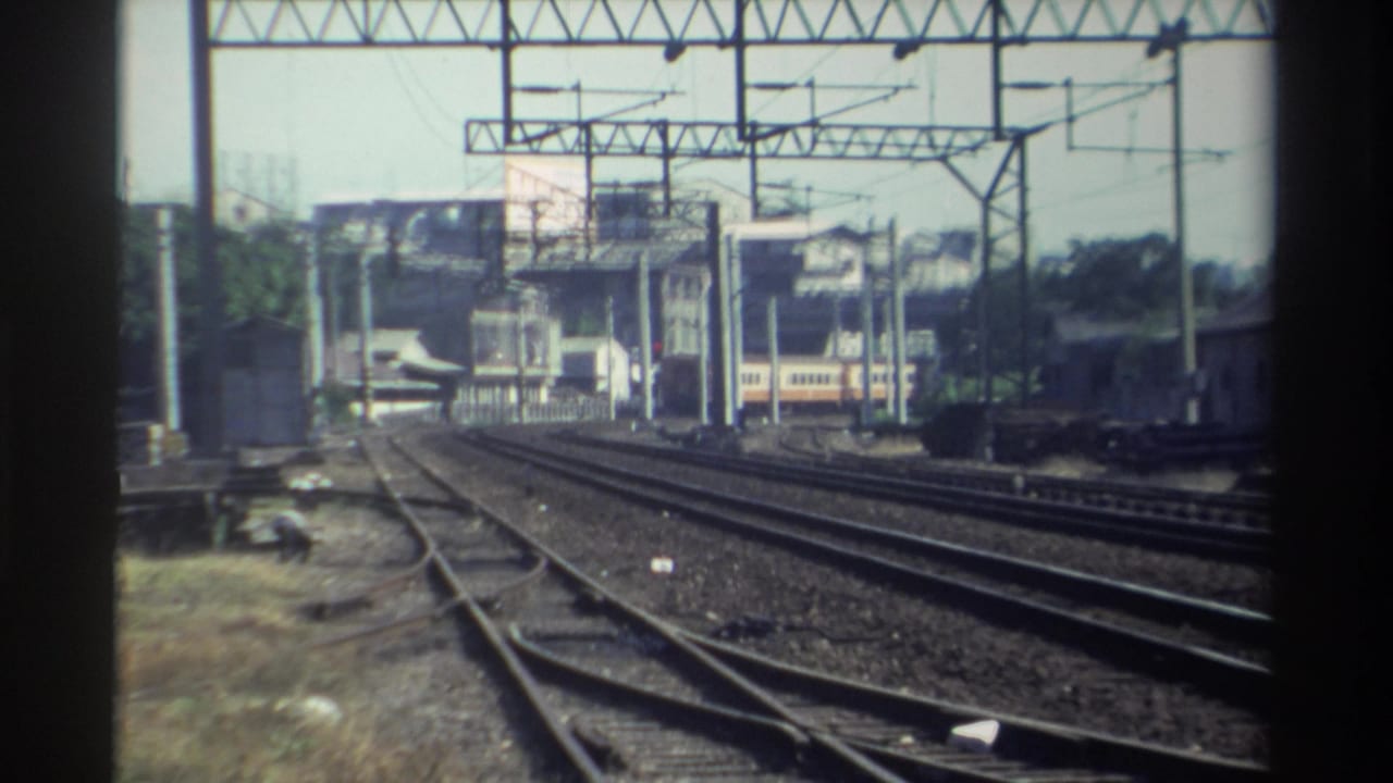 Railroad tracks and overhead wire supports lead toward a small train near buildings and trees.