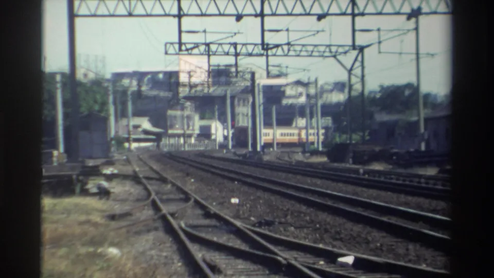1982 Taiwan rail scene with converging tracks, overhead catenary supports, a distant train, trees, and surrounding low-rise structures.