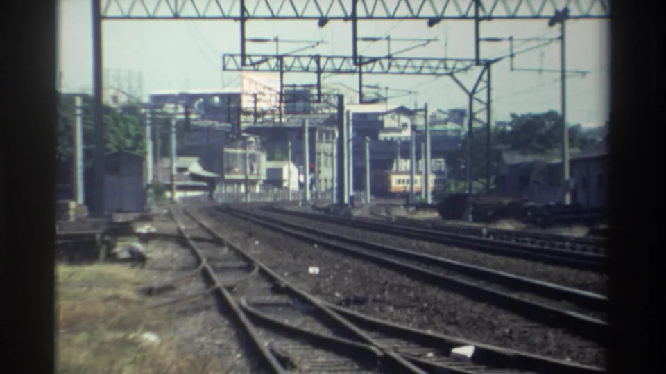1982 Taiwan 8mm frame showing a broad rail corridor, overhead electric supports, a distant train, and rail-side buildings under bright sky.