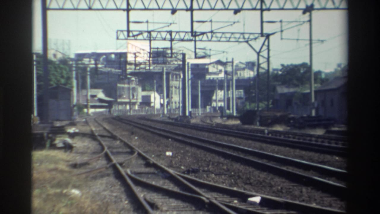 Rail corridor with multiple tracks, overhead wires, and buildings near a station area.