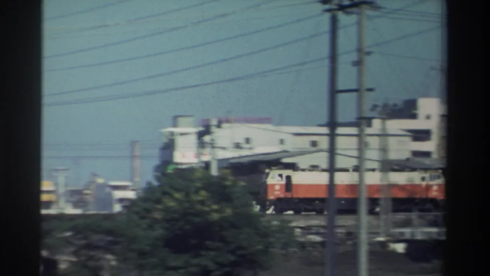 1982 Taiwan frame with a red-and-white locomotive moving past utility poles, wires, trees, and low-rise buildings.