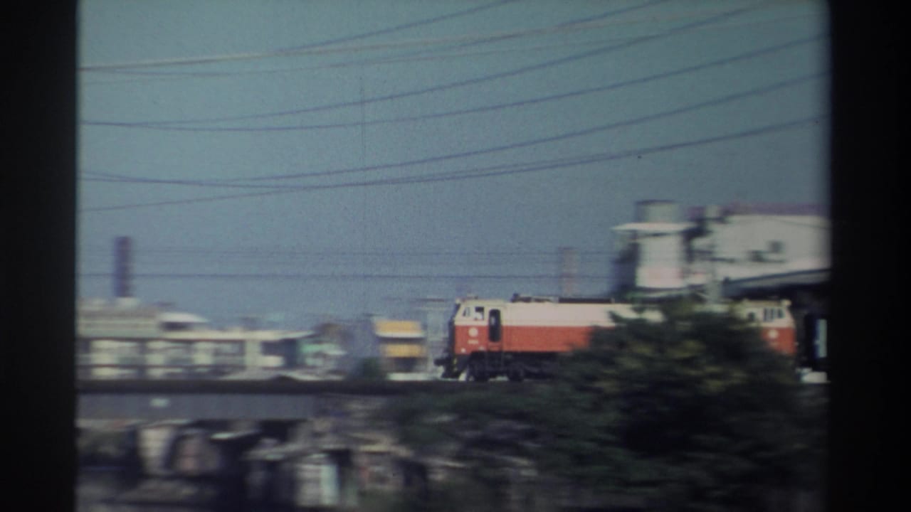 1982 Taiwan 8mm frame showing a red-and-white locomotive moving across a concrete river bridge, with utility lines and low industrial buildings in the background.