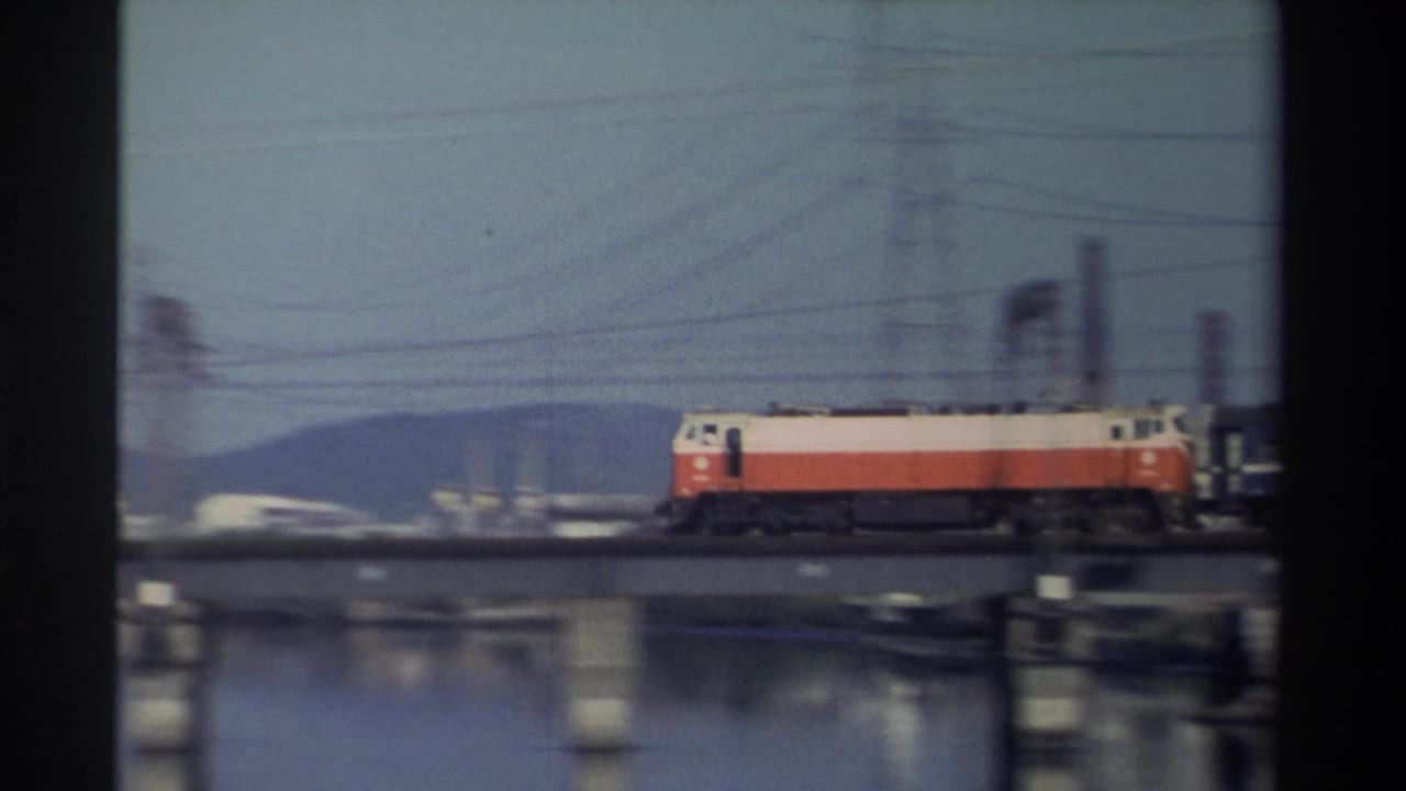 1982 Taiwan 8mm frame of a red-and-white locomotive crossing left to right over a river bridge, with utility infrastructure and distant hills visible.