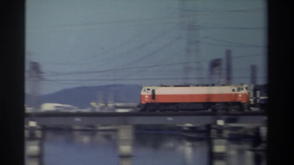 1982 Taiwan 8mm frame of a red-and-white locomotive crossing left to right over a river bridge, with utility infrastructure and distant hills visible.