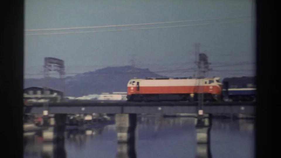 1982 Taiwan home-movie frame showing the locomotive pulling a dark passenger car across a concrete bridge over water, with low buildings and hills beyond.