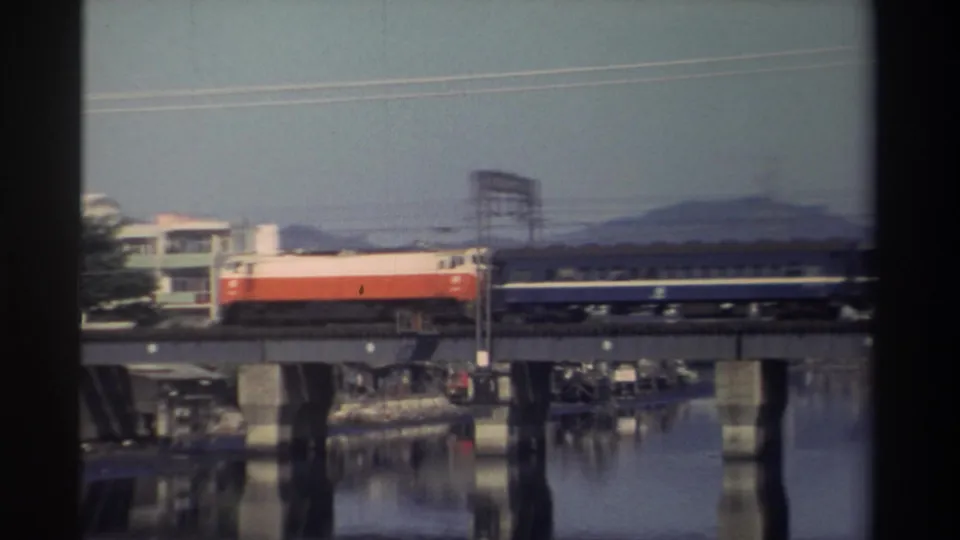 1982 Taiwan 8mm frame of a red-and-white locomotive pulling a passenger car across a river bridge, with reflections, buildings, and distant hills visible.