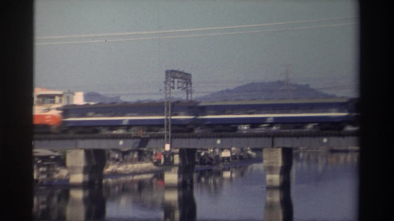 1982 Taiwan home-movie frame showing a passenger train moving across a concrete river bridge with hills and power lines behind it.