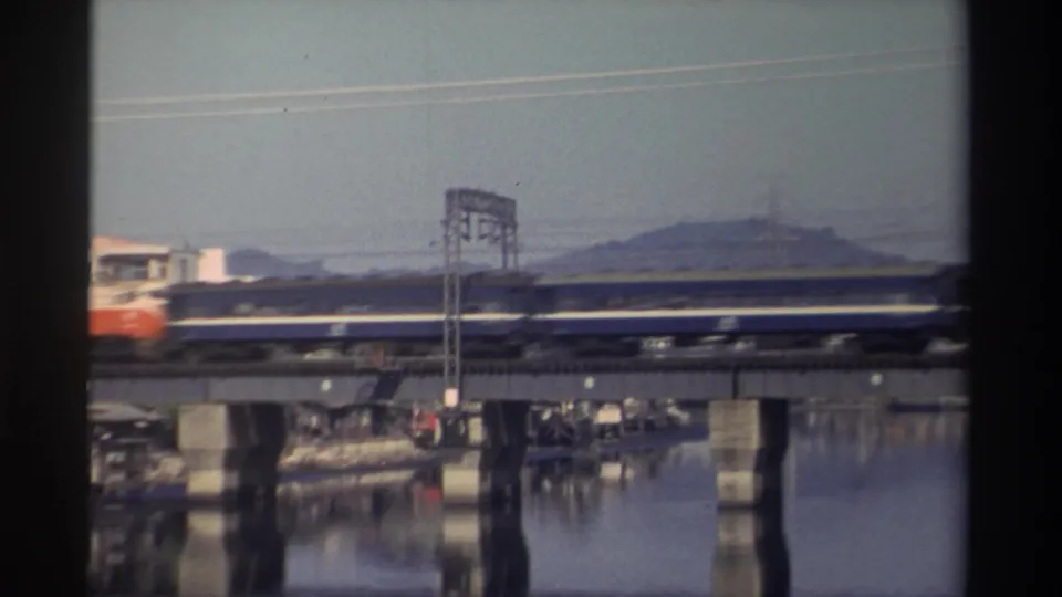 1982 Taiwan home-movie frame showing a passenger train moving across a concrete river bridge with hills and power lines behind it.