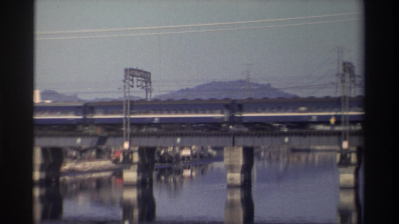 The train passes left to right across a concrete bridge in Taiwan, with power structures, still water, and a hazy hillside beyond.