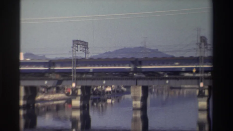 The train passes left to right across a concrete bridge in Taiwan, with power structures, still water, and a hazy hillside beyond.