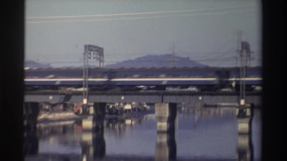 A soft 8mm frame shows the blue-and-white train midway across the river bridge, backed by overhead wires and a low hill.