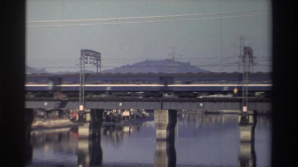 The passenger train nears the right edge of the bridge while calm water, concrete piers, power lines, and distant hills remain steady.