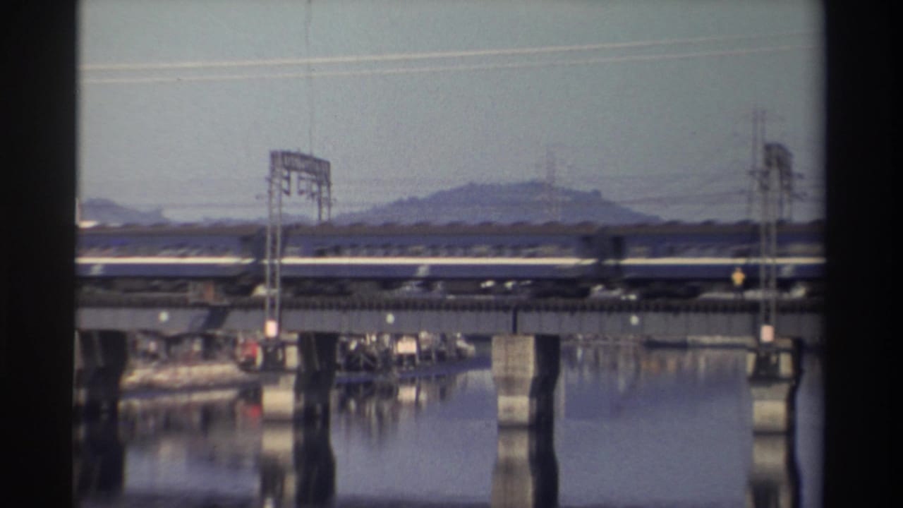 Blue passenger train crossing a concrete bridge over a calm river with a hill behind it.