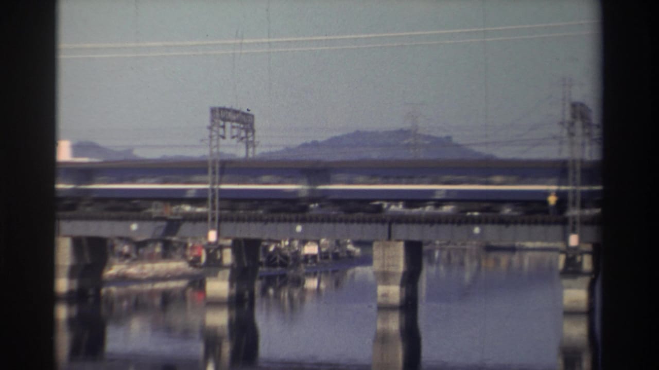 Passenger train on a bridge above still water, with utility towers and a hill in the background.