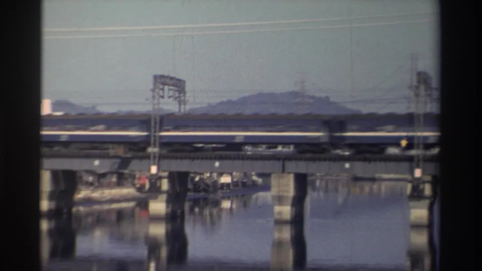 1982 Taiwan 8mm frame with the passenger train mid-crossing, backed by a hill, utility lines, river reflections, and small structures near the water.