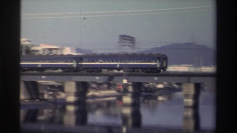 1982 Taiwan frame showing the rear cars of a blue passenger train on a concrete river bridge, with blurred motion and hills beyond.