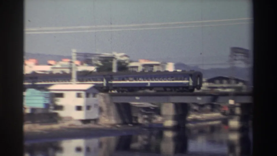 1982 Taiwan 8mm frame showing a blue passenger train moving across a bridge above a waterfront with buildings and river reflections.