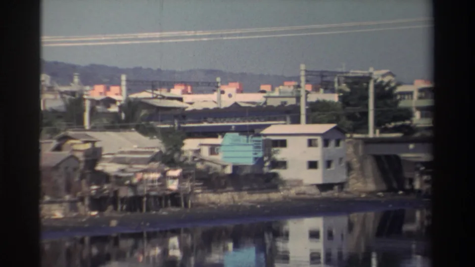 1982 Taiwan waterfront view with low-rise buildings, bridge supports, utility poles, and calm river reflections after the train has passed.