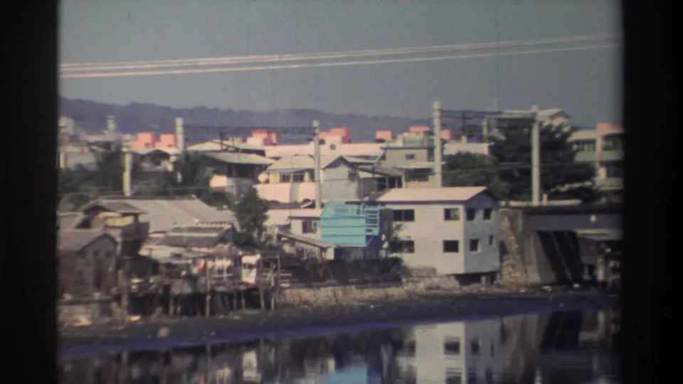 1982 Taiwan 8mm frame showing riverside houses, utility poles, a bridge edge, and still water reflecting the waterfront.