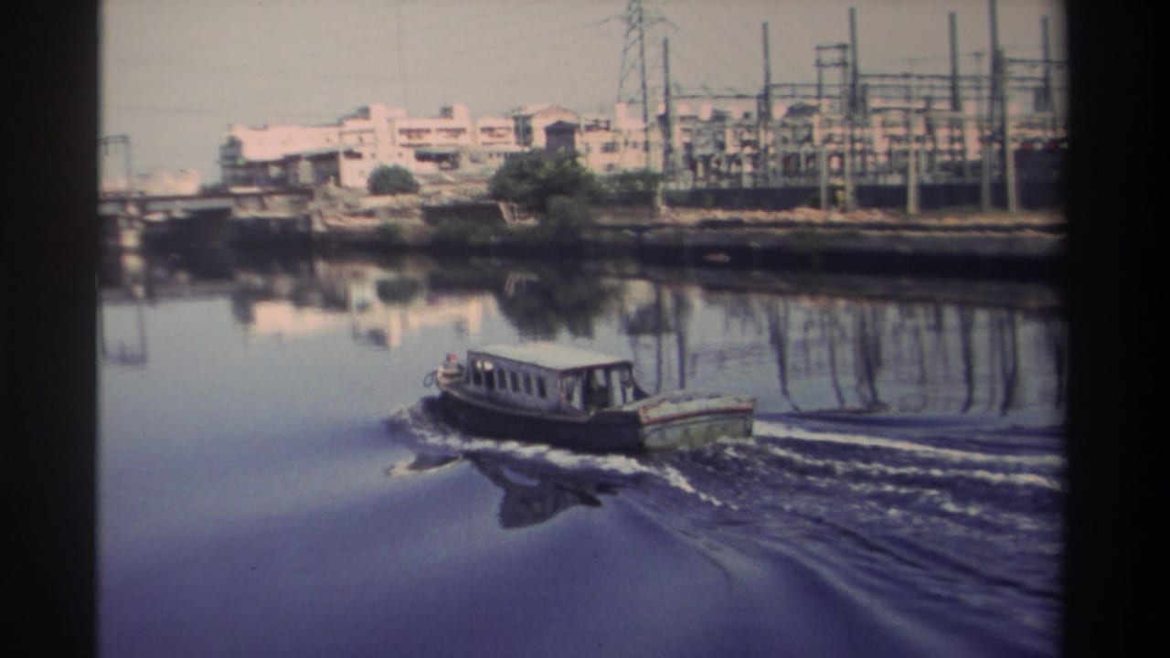 1982 Taiwan 8mm frame of a small motorboat leaving a wake on a calm urban river near bridge spans and utility infrastructure.