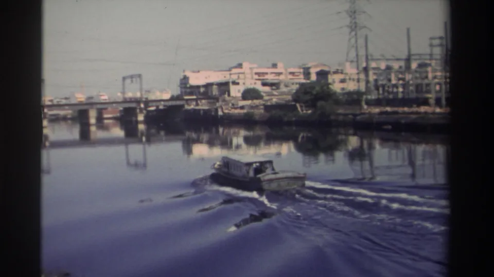 1982 Taiwan 8mm frame showing a small river boat receding along an urban waterway with bridge spans and shoreline infrastructure.
