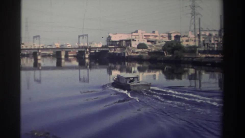 1982 Taiwan home-movie frame of a small motorboat farther downriver, with bridge spans, power lines, and city shoreline behind it.