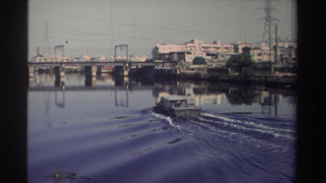1982 Taiwan 8mm frame showing a small tugboat leaving a wake on river water near bridge supports and industrial buildings.