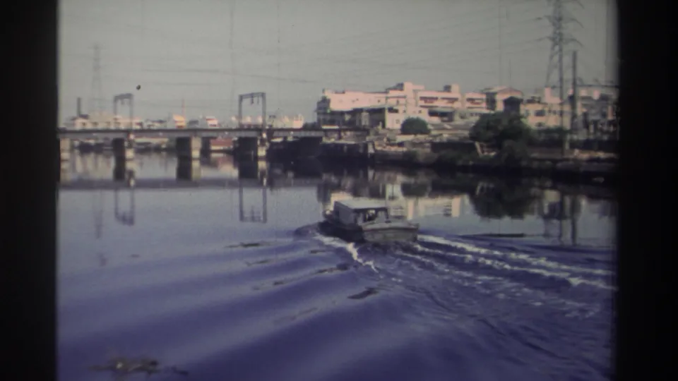 1982 Taiwan 8mm frame showing a small tugboat leaving a wake on river water near bridge supports and industrial buildings.