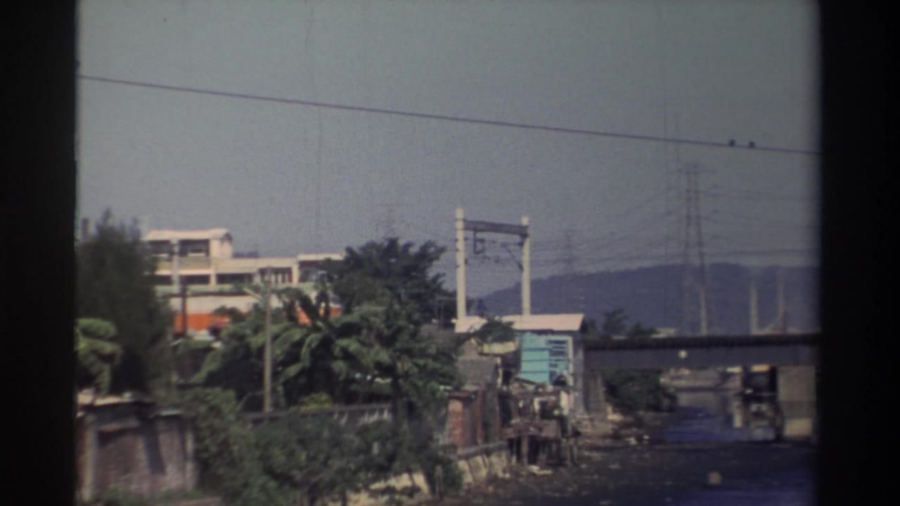 1982 Taiwan frame with an orange-and-white train crossing a bridge above a riverbank, power lines, and distant hills.