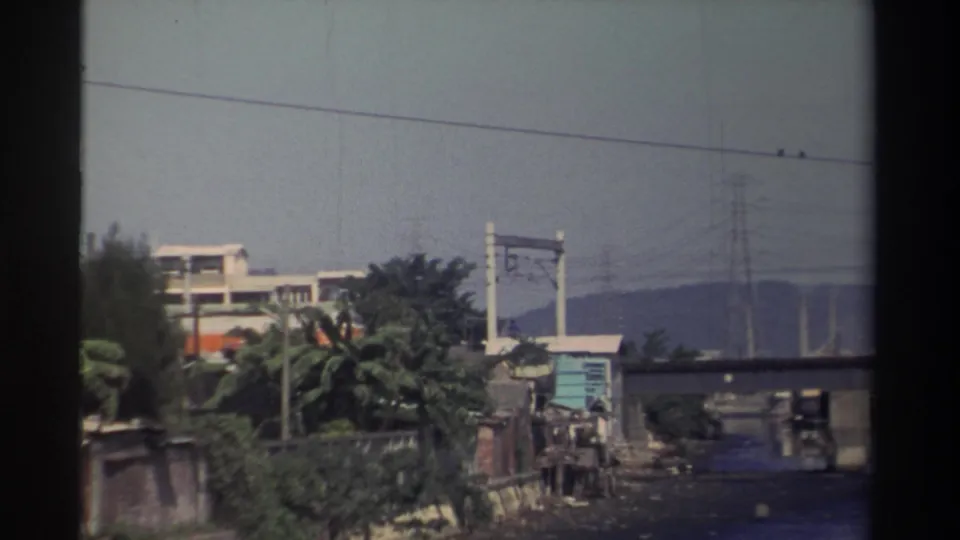 1982 Taiwan frame with an orange-and-white train crossing a bridge above a riverbank, power lines, and distant hills.