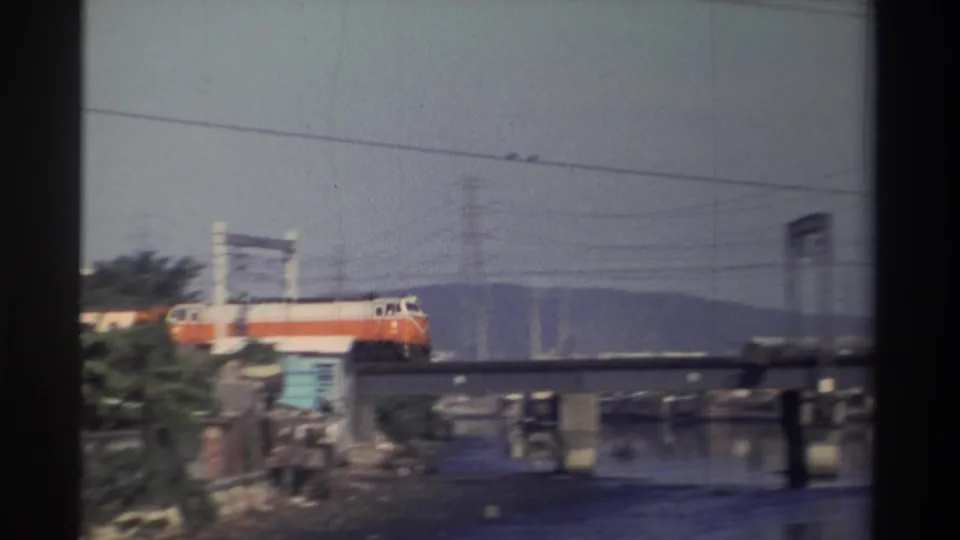 1982 Taiwan 8mm view of an orange train moving across a river bridge, partly hidden by trees and foreground structures.