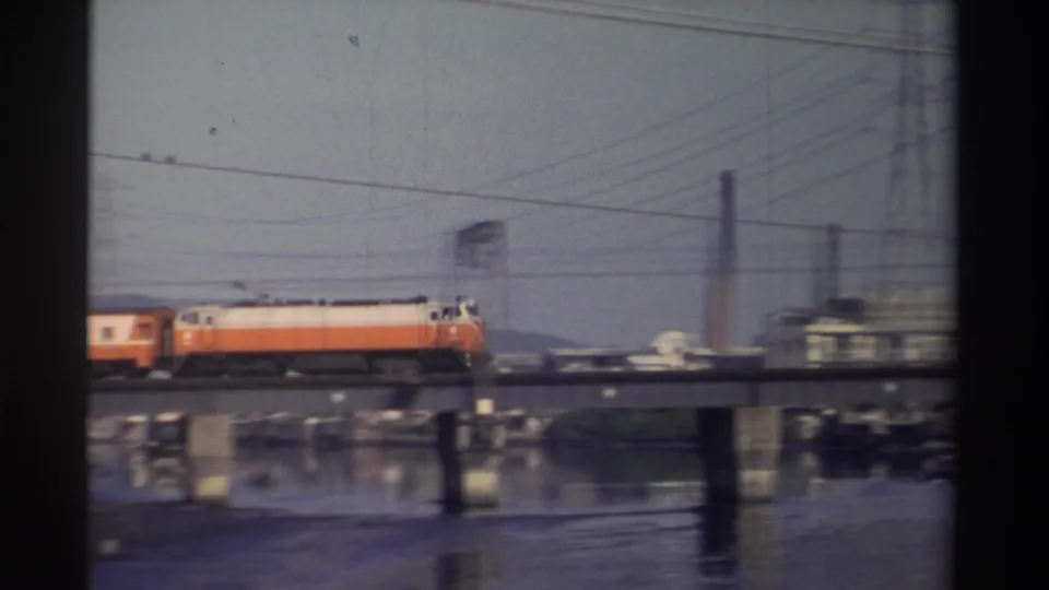 1982 Taiwan frame showing an orange-and-white train on a bridge over river water, with pylons, wires, and industrial structures nearby.