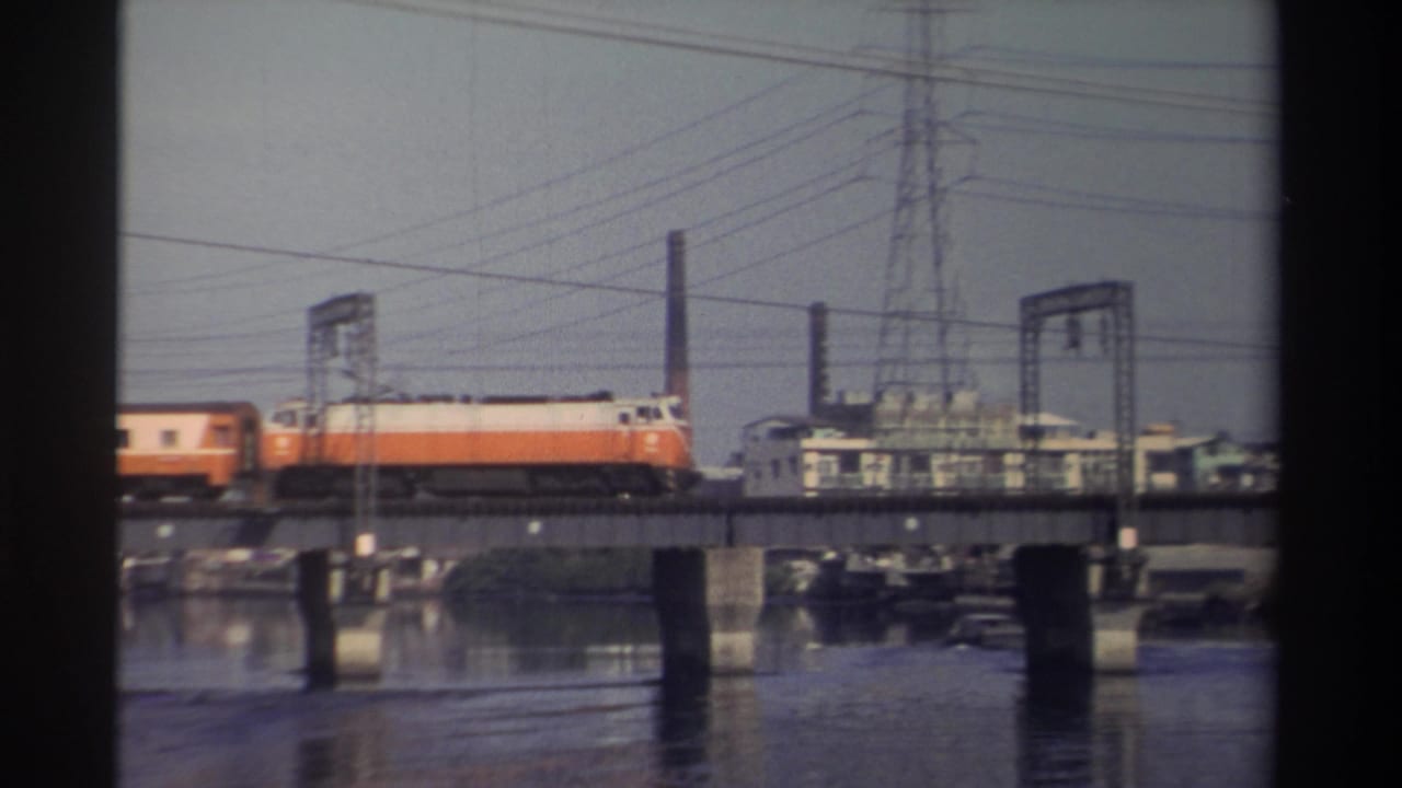 1982 Taiwan home-movie frame showing an orange-and-white train moving right across a bridge over water, with wires and industrial buildings in the background.