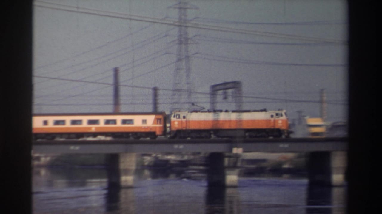 Passenger car and orange-and-white locomotive crossing a bridge with power lines overhead.