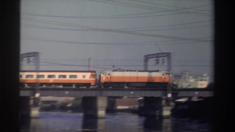A soft 8mm frame shows the orange-and-white train crossing a concrete bridge over water, with utility lines and industrial structures behind it.