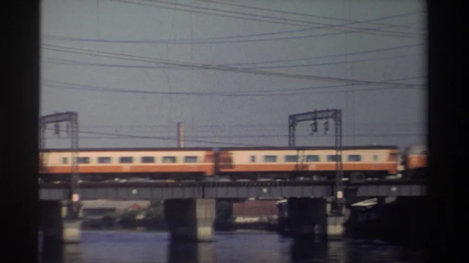 Two orange-and-white passenger cars move right across the bridge in the final frame, with water, piers, utility wires, and industrial structures visible.