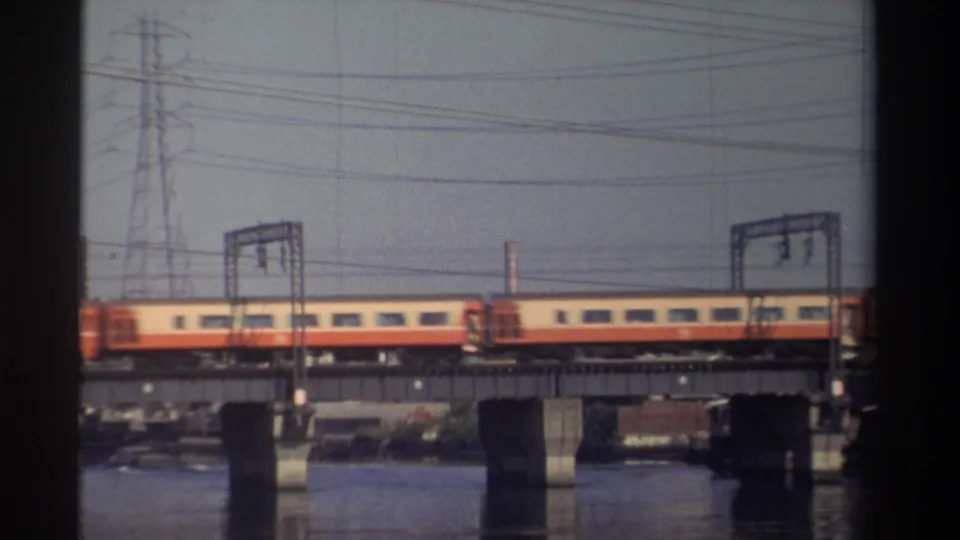 Passenger train continues across a bridge over water in Taiwan, with electrical gantries and a utility tower visible.
