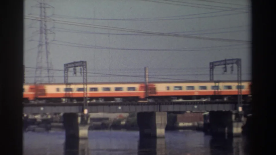 Soft 8mm frame of a passenger train crossing a waterway bridge, with rail gantries and cars below or beyond.