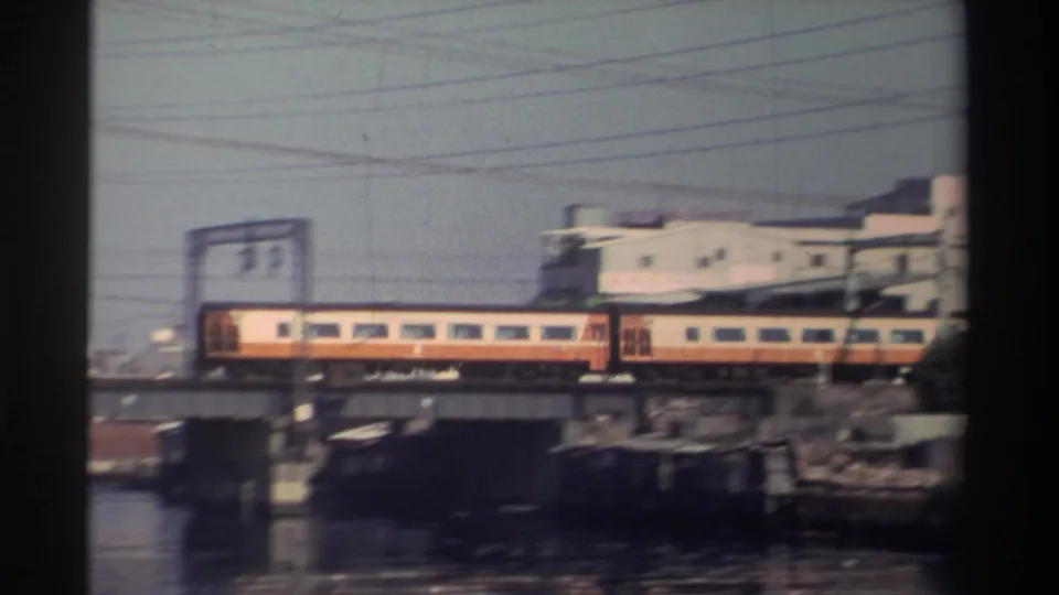 1982 Taiwan 8mm frame showing a cream-and-orange train crossing a river bridge beside low-rise industrial buildings and utility wires.