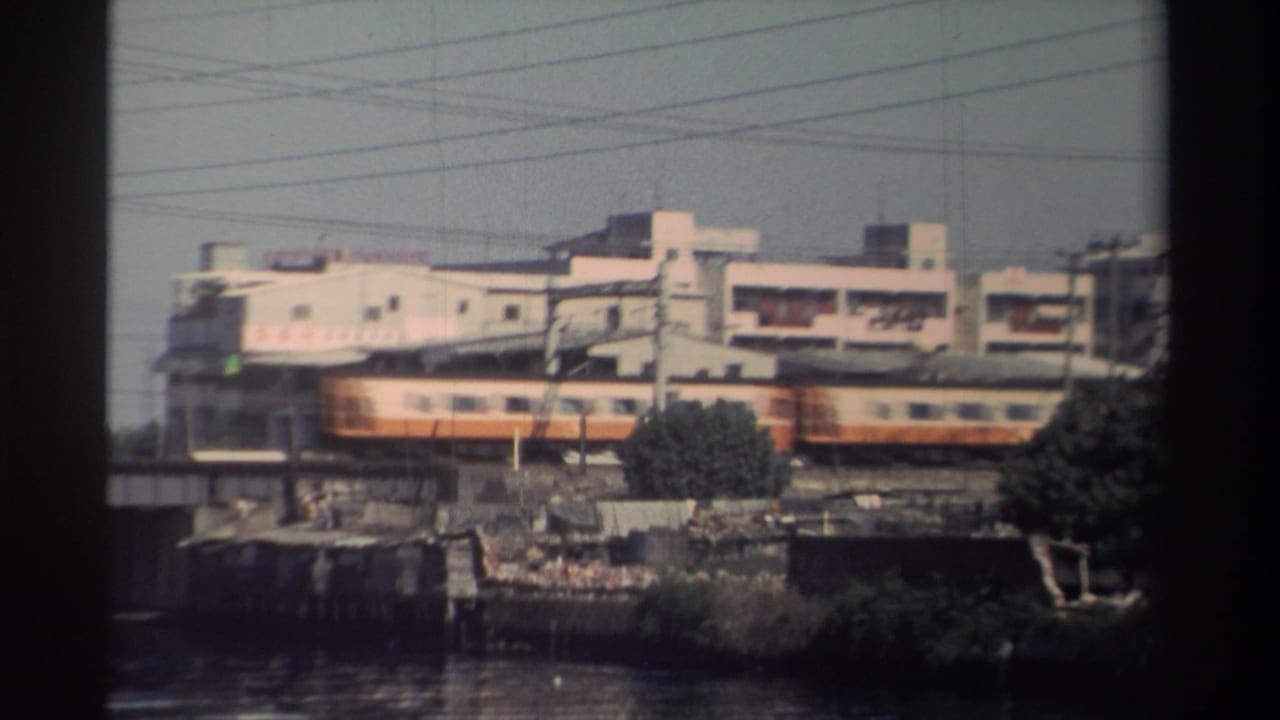 1982 Taiwan 8mm frame with train cars moving past industrial buildings, utility poles, overhead wires, and a riverbank tree.
