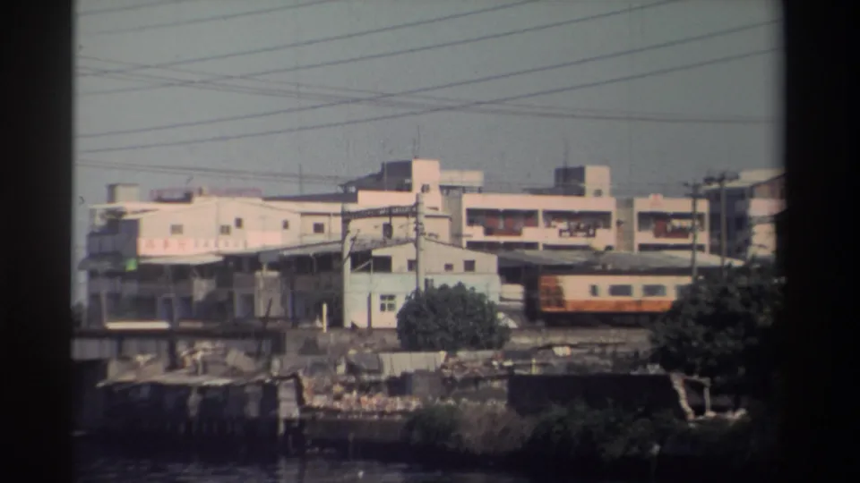 1982 Taiwan 8mm frame showing the rear of a cream-and-orange train crossing near industrial buildings, overhead wires, and riverside trees.