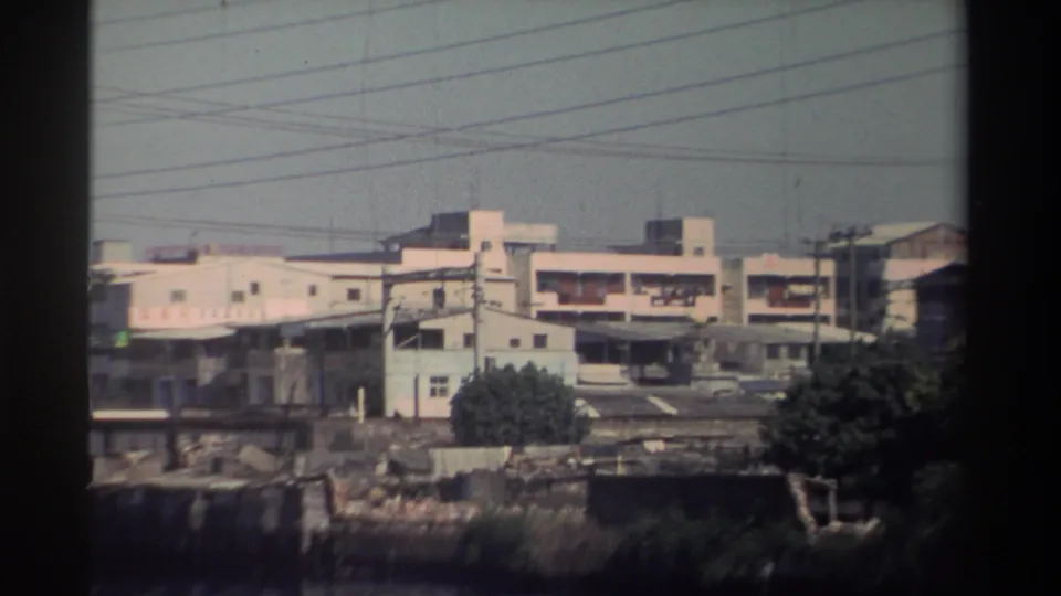 1982 Taiwan 8mm frame where the train has mostly left view, leaving industrial buildings, utility wires, bridgework, and riverside vegetation.