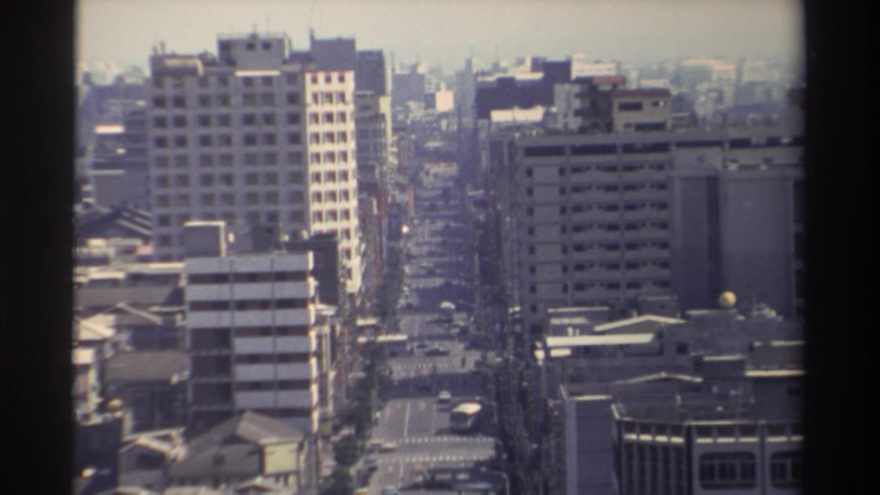 High-angle 1982 Taiwan street view with modern blocks, rooftops, light traffic, and a soft hazy skyline.