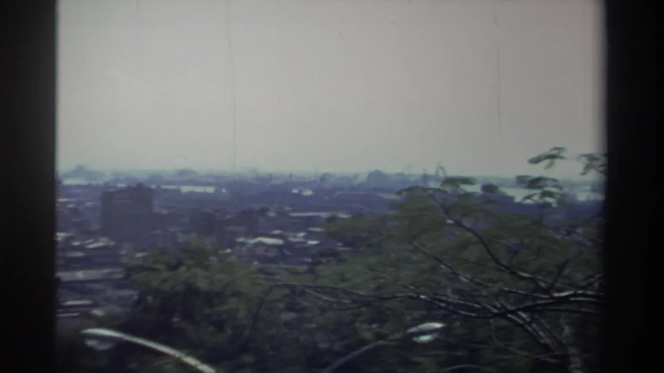 1982 Taiwan home-movie frame looking over branches toward a hazy cityscape, a taller building, and possible water in the distance.