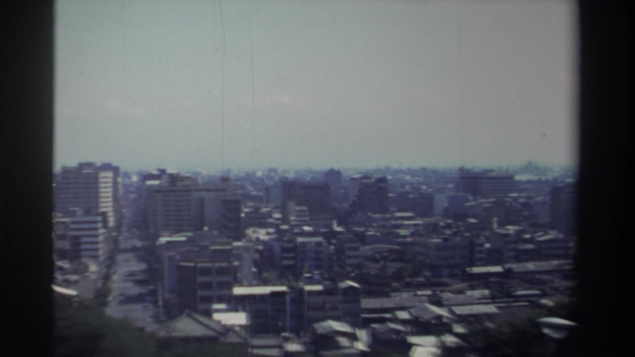 1982 Taiwan home-movie frame showing a soft, elevated city panorama with mid-rise blocks, narrow streets, foreground trees, and dark film edges.