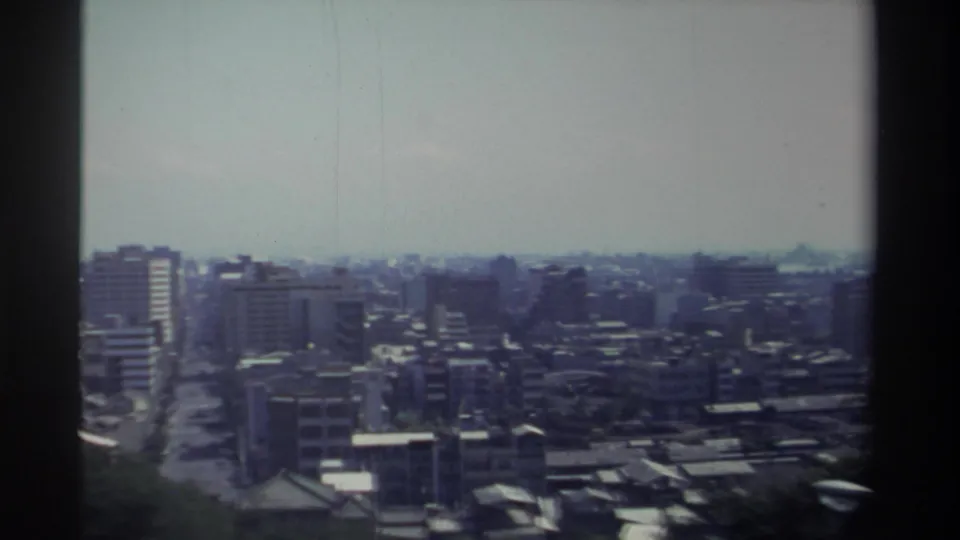 1982 Taiwan home-movie frame showing a soft, elevated city panorama with mid-rise blocks, narrow streets, foreground trees, and dark film edges.