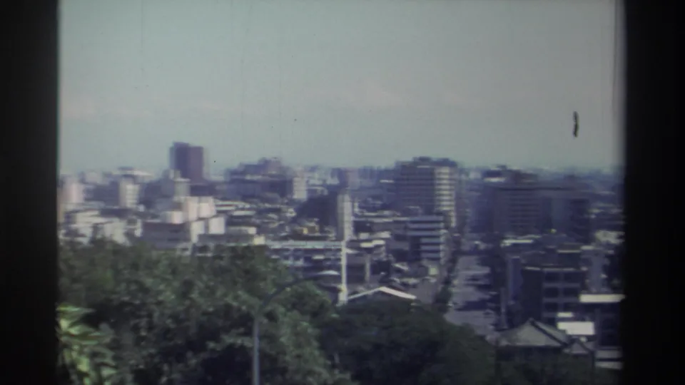 Elevated 1982 Taiwan city view with a darker tower at left, clustered mid-rises, a central street, trees, and visible 8mm softness.