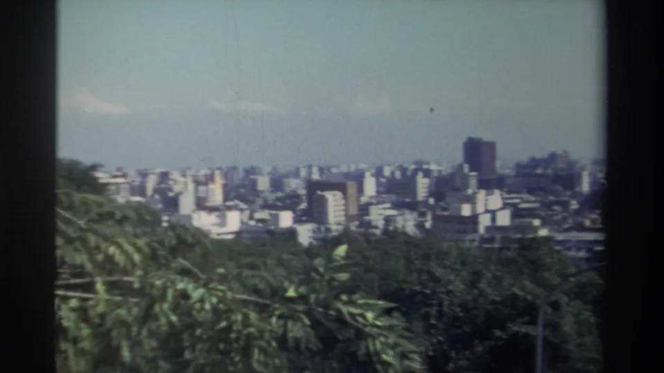 1982 Taiwan home-movie frame showing a high-angle city view framed by green trees, with dense buildings and a taller tower in the distance.